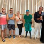 Un retrato de grupo de los seis sospechosos clave para una actividad de team building de Cluedo corporativo. Cada empleado lleva un disfraz distintivo que representa un personaje: un invitado informal, un monitor deportivo, una conserje de hotel con placa de identificación, un jardinero hosco con rastrillo y botas de agua, un hombre de negocios elegante y una viuda dramática vestida de negro con tocado y perlas. Posan en fila en un moderno vestíbulo de hotel, listos para que los equipos los interroguen.