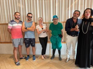 Un retrato de grupo de los seis sospechosos clave para una actividad de team building de Cluedo corporativo. Cada empleado lleva un disfraz distintivo que representa un personaje: un invitado informal, un monitor deportivo, una conserje de hotel con placa de identificación, un jardinero hosco con rastrillo y botas de agua, un hombre de negocios elegante y una viuda dramática vestida de negro con tocado y perlas. Posan en fila en un moderno vestíbulo de hotel, listos para que los equipos los interroguen.